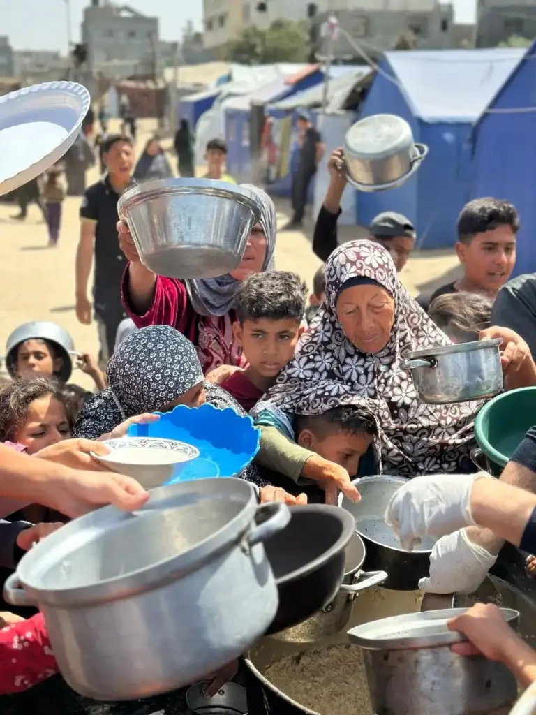Displaced families in Gaza holding pots and bowls in line to receive food from Rammun Foundation